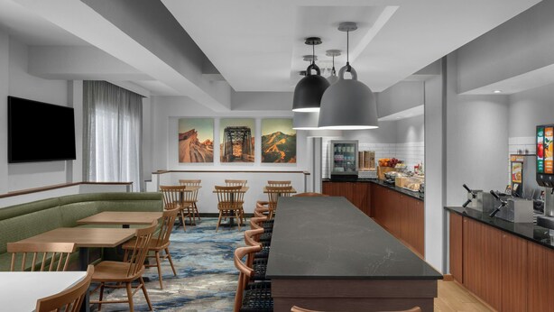 Uma área de refeições moderna em hotel, com uma mesa comunitária grande, cadeiras de madeira, um assento estofado verde e um balcão de cozinha.