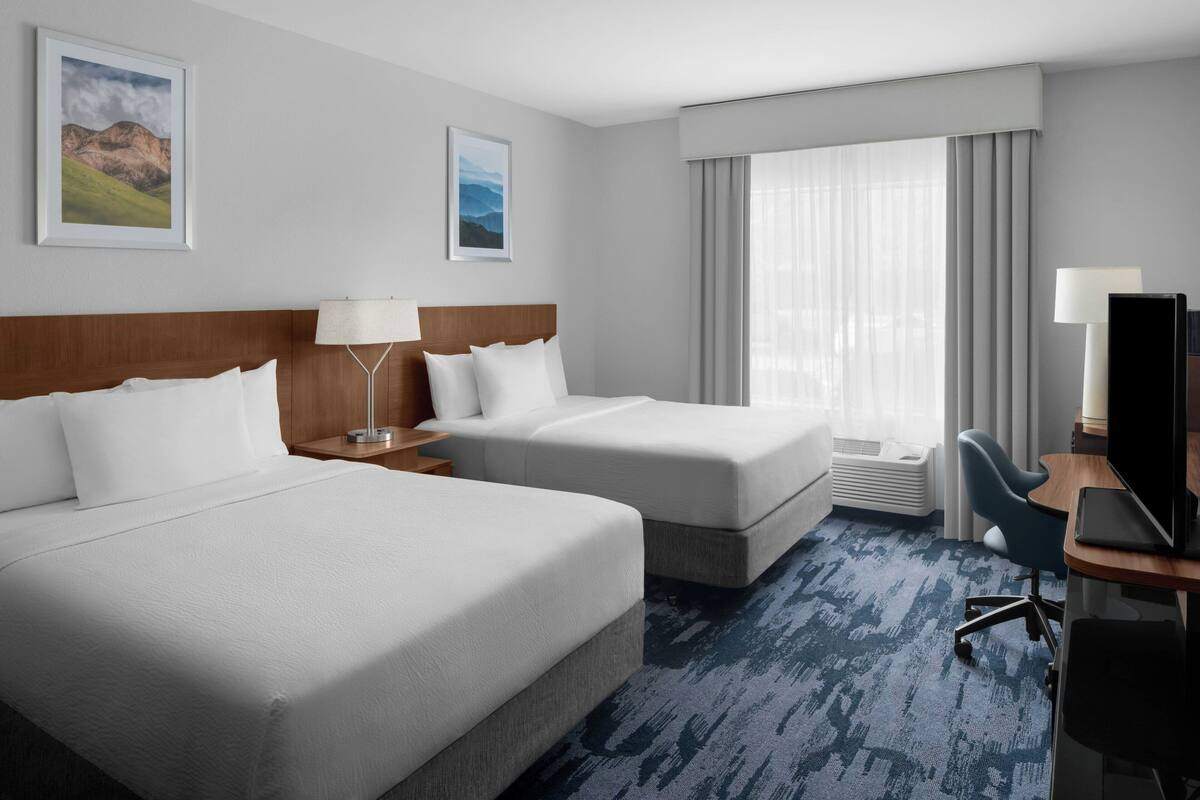A hotel room with two beds, a desk with a computer, a chair, and a large window with curtains.