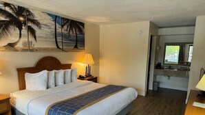A hotel room with a bed, a nightstand, a lamp, a painting of palm trees, and a view of the kitchen. - Days Inn by Wyndham Chiefland (Chiefland)