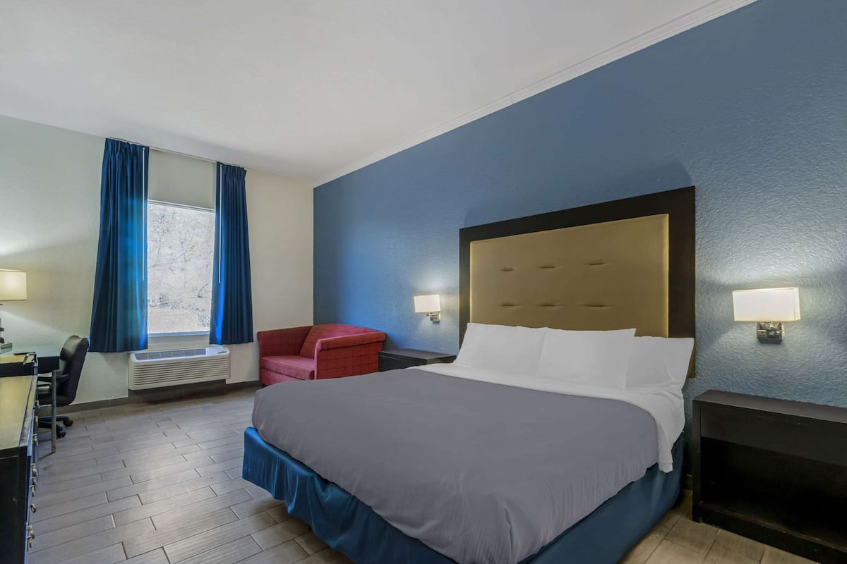A hotel room with a large bed, a desk with a chair, a red armchair, a window with blue curtains, and a wall-mounted lamp.