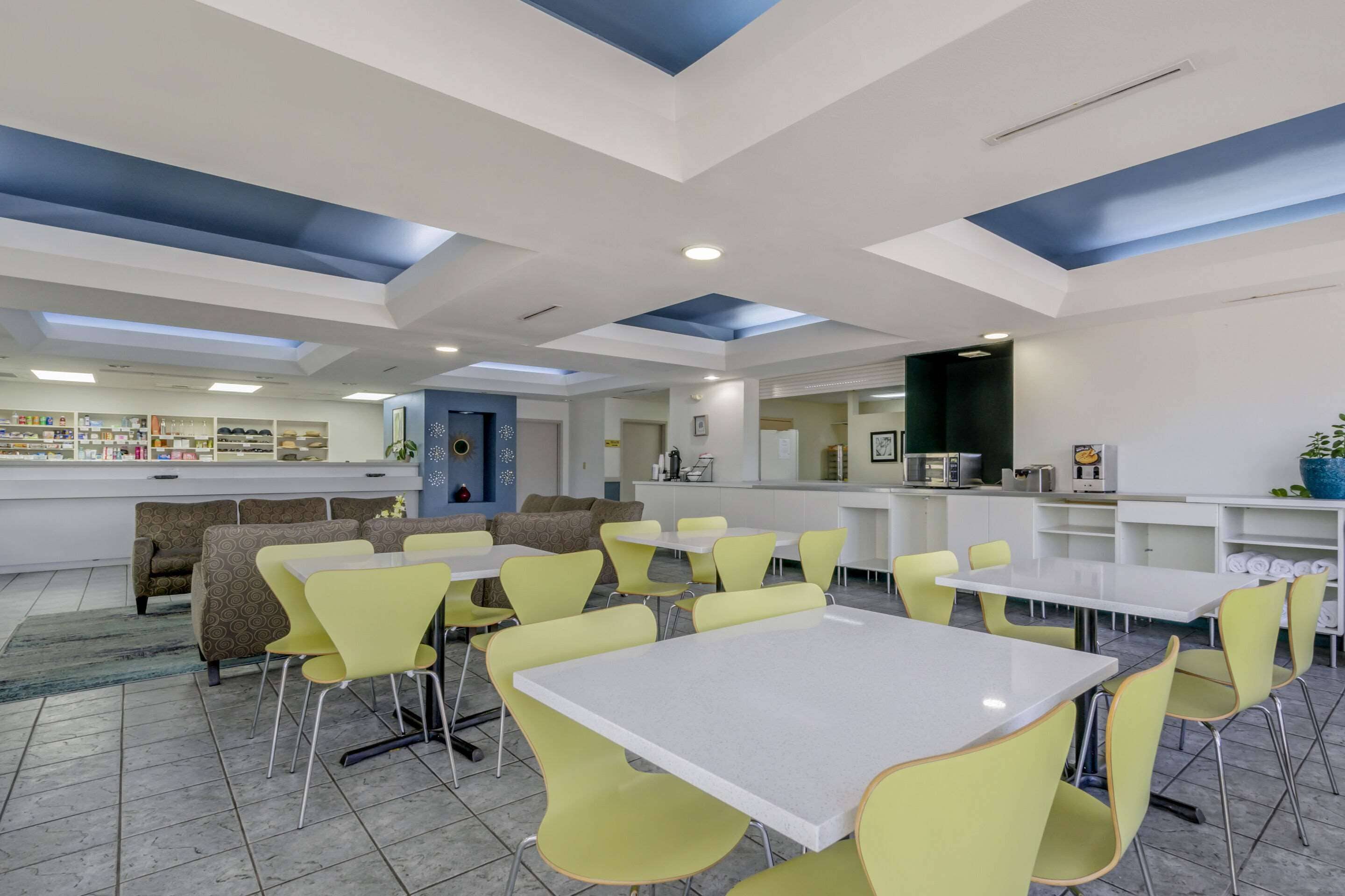 A modern dining area with yellow chairs and white tables, a bar area with shelves, and a seating area with patterned sofas.