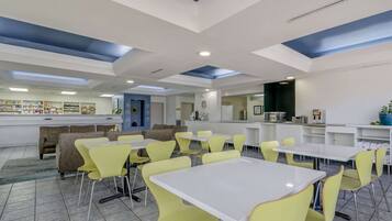 A modern dining area with yellow chairs and white tables, a bar area with shelves, and a seating area with patterned sofas.