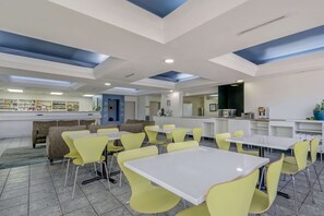 A modern dining area with yellow chairs and white tables, a bar area with shelves, and a seating area with patterned sofas.