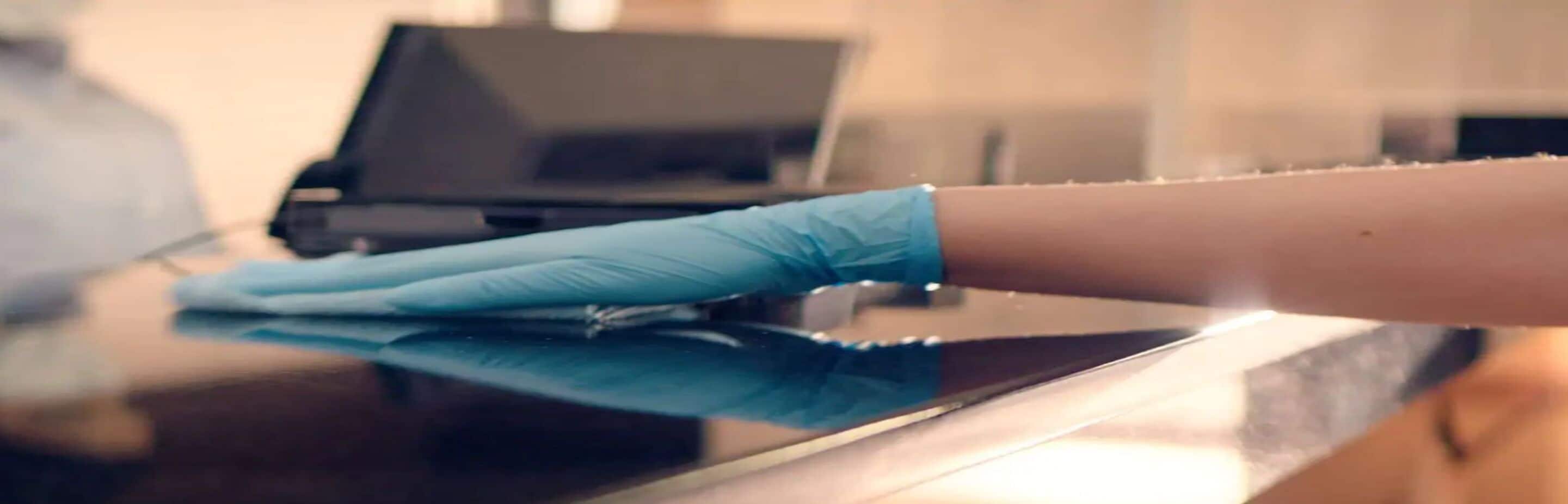 Une main portant un gant bleu nettoie une surface réfléchissante, probablement une table ou un comptoir.