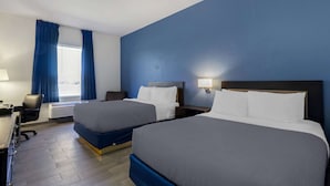 A hotel room with two beds, a desk, a chair, a window with curtains, and a wall-mounted lamp. - Rodeway Inn Wesley Chapel (Wesley Chapel)