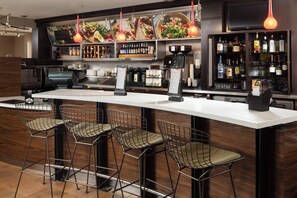 A bar area with a long counter, multiple stools, a coffee machine, and a variety of liquor bottles. - Courtyard by Marriott Indianapolis at the Capitol (Indianapolis)