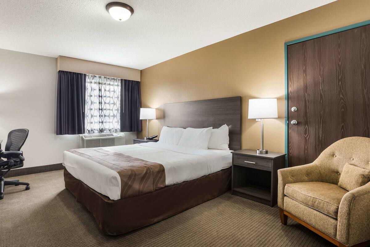 Una habitación de hotel con una cama grande, dos lámparas de noche, una silla, un escritorio y una ventana con cortinas.