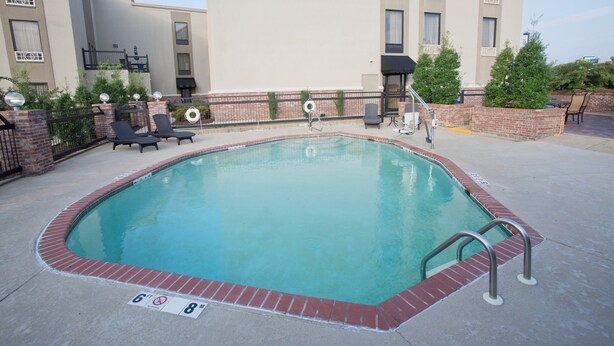 A rectangular swimming pool with a brick edge, surrounded by a brick wall and a metal fence. There are lounge chairs and a barbecue grill visible.