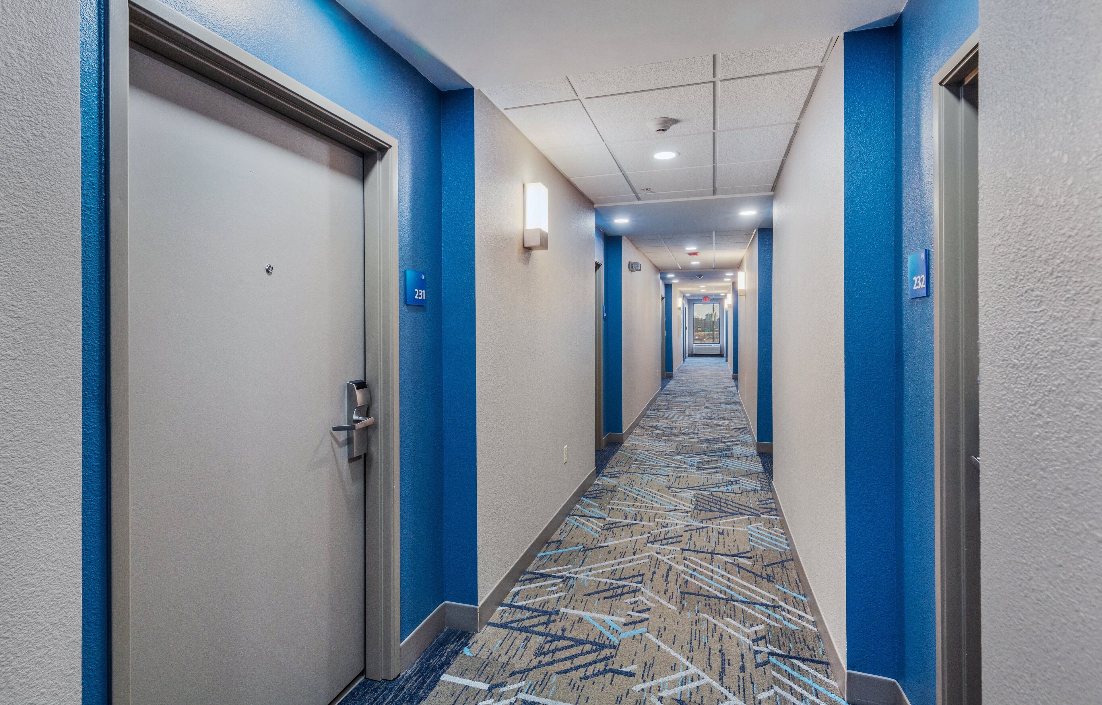 A hotel hallway with blue and white walls, numbered doors, and a patterned carpet.