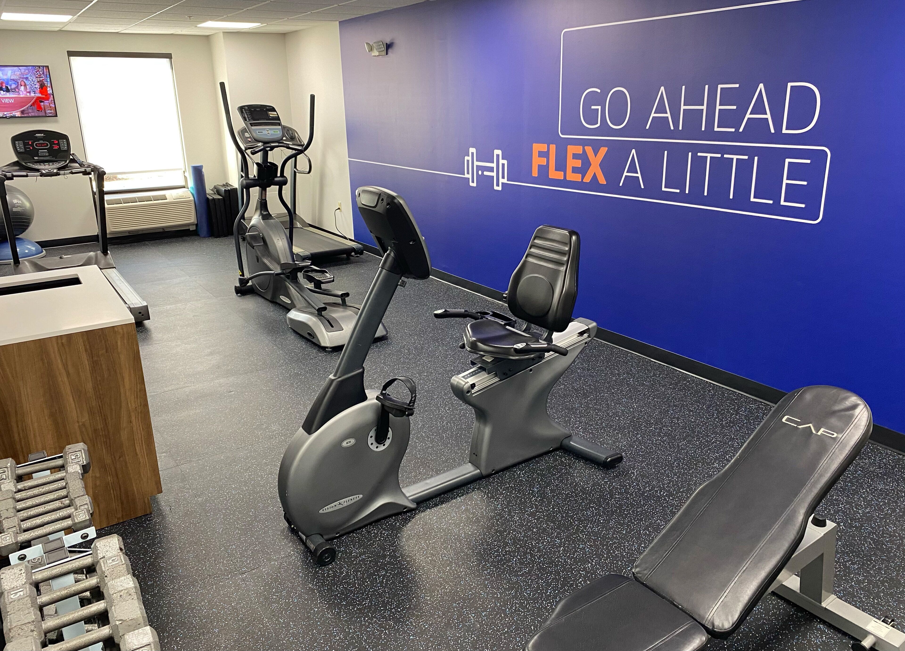 A gym with cardio machines, dumbbells, and a wall with motivational text.