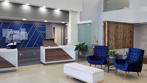 A hotel reception area with a modern design, featuring a reception desk, blue upholstered chairs, and a white bench. - Holiday Inn Express New Albany by IHG (New Albany)