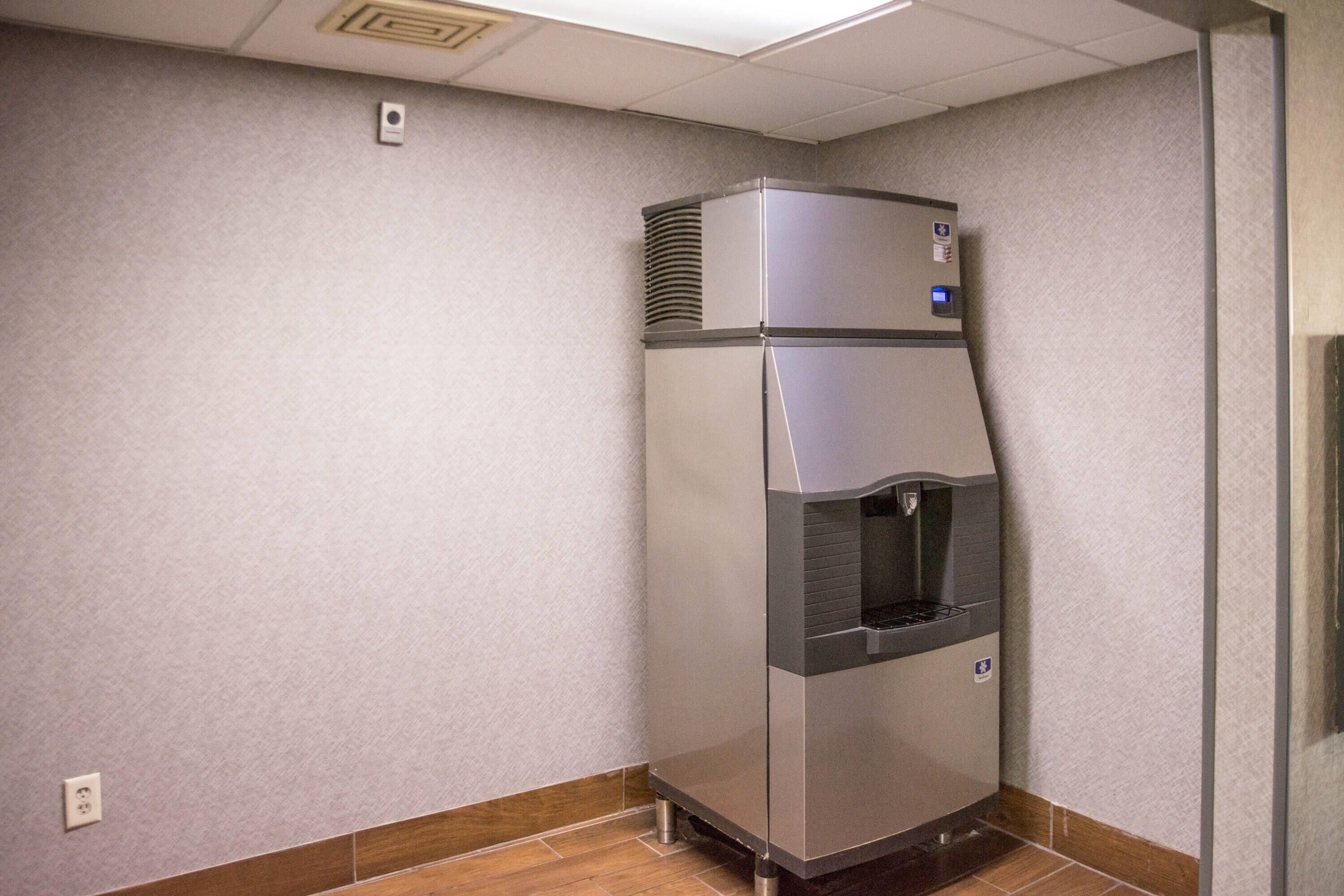 A compact, stainless steel refrigerator with a water dispenser, located in a corner of a room with a tiled floor and a textured wall.