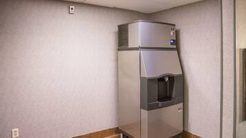 A compact, stainless steel refrigerator with a water dispenser, located in a corner of a room with a tiled floor and a textured wall.