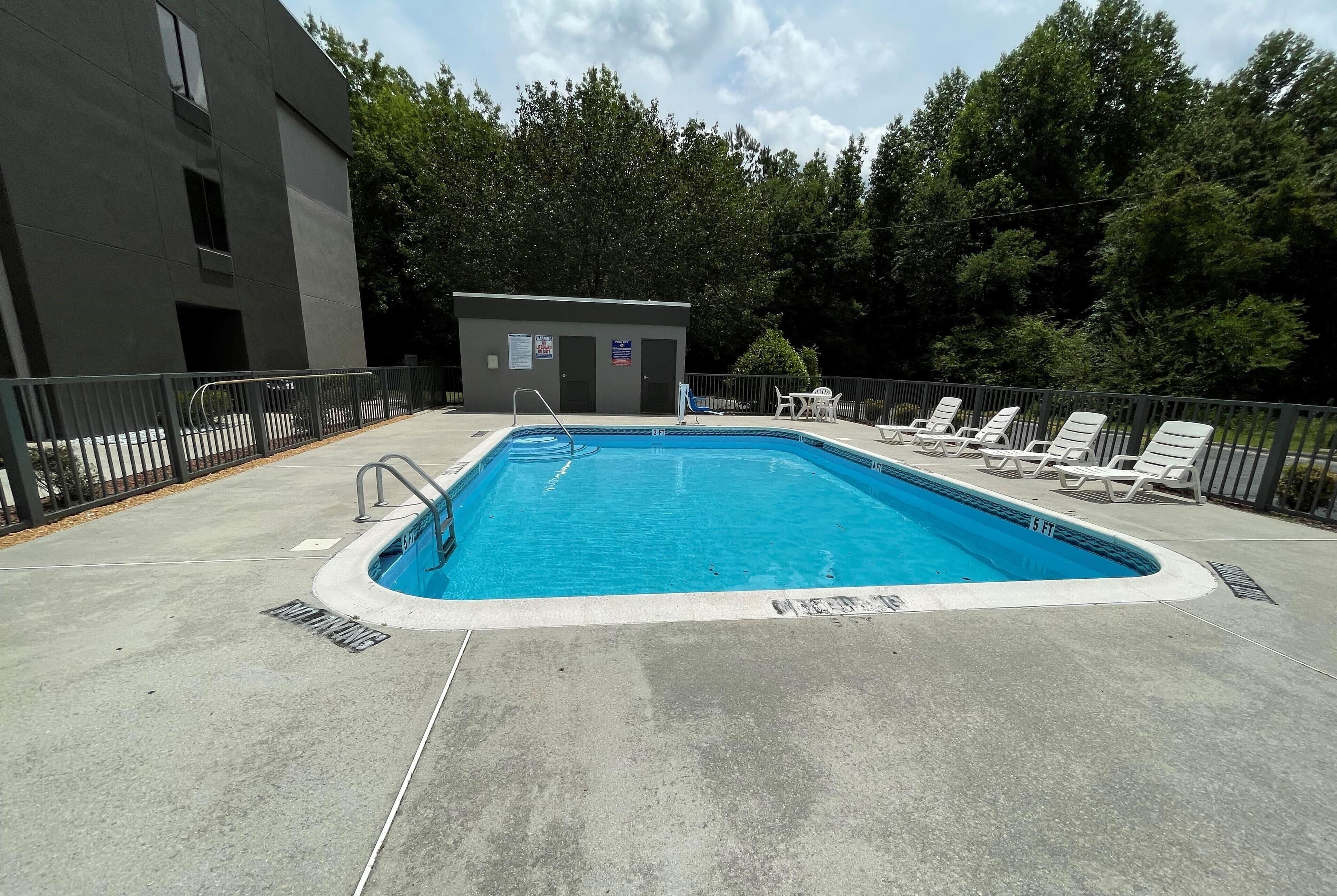 A rectangular swimming pool with a metal ladder, surrounded by a concrete deck. There are lounge chairs and a small building with a door and windows.