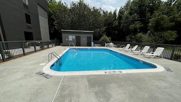Una piscina rectangular con una escalera metálica, rodeada por una plataforma de concreto. Hay sillas de descanso y un pequeño edificio con una puerta y ventanas.
