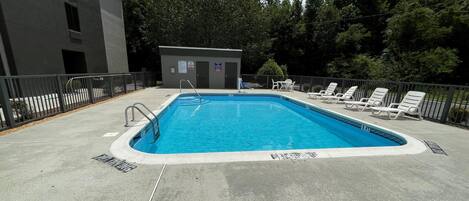 Una piscina rectangular con una escalera metálica, rodeada por una plataforma de concreto. Hay sillas de descanso y un pequeño edificio con una puerta y ventanas.