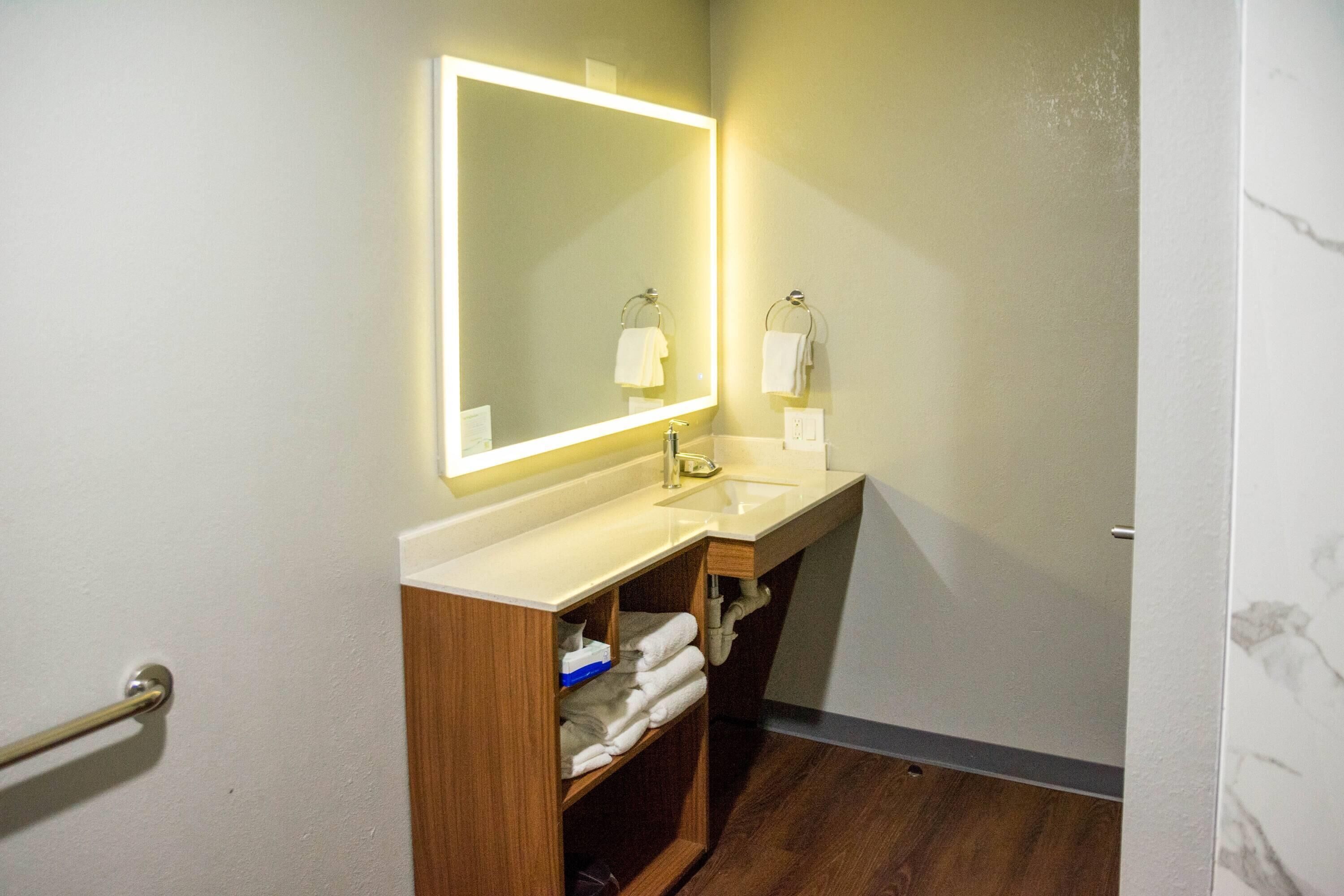 Un baño con tocador de madera, un espejo grande con iluminación debajo del mueble y un perchero para toallas.