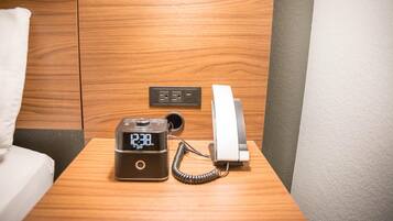 Una mesita de noche de madera con un reloj digital, un teléfono y una toma de corriente.