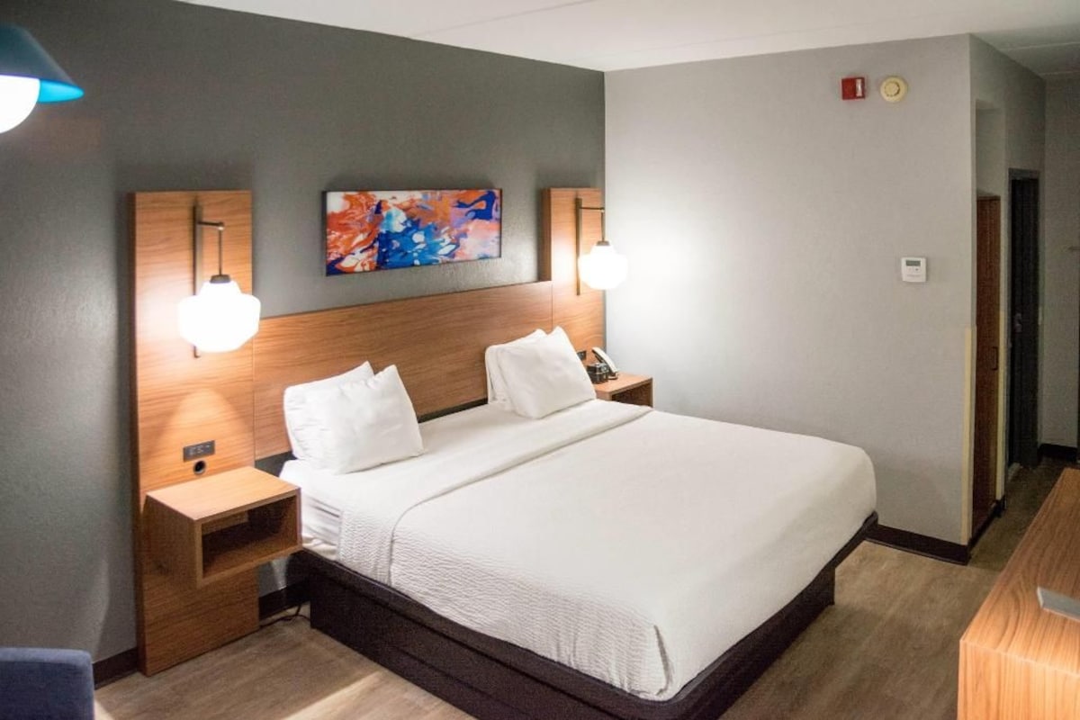A hotel room with a large bed, two bedside tables, a headboard with artwork, and wall-mounted lamps.