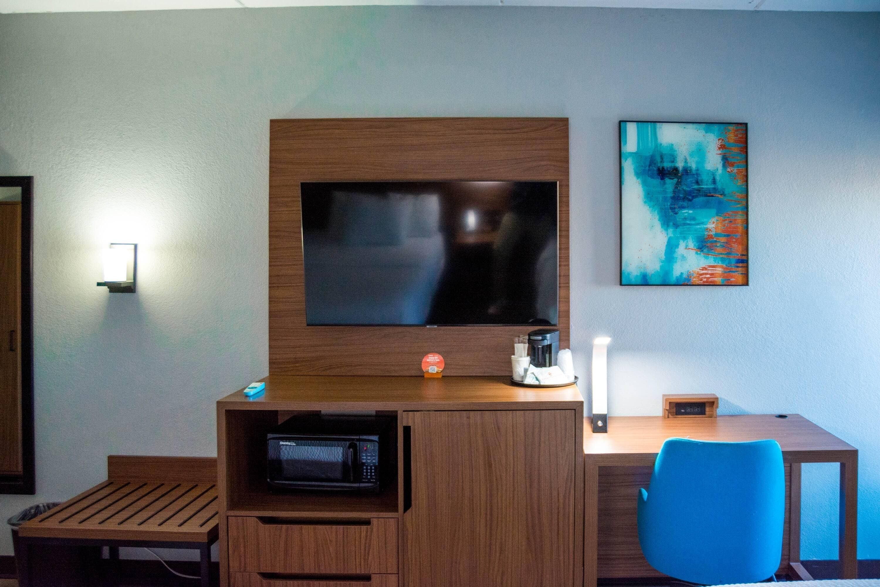 A hotel room with a wooden desk, a blue chair, a flat-screen TV mounted on the wall, a microwave, and a coffee maker.