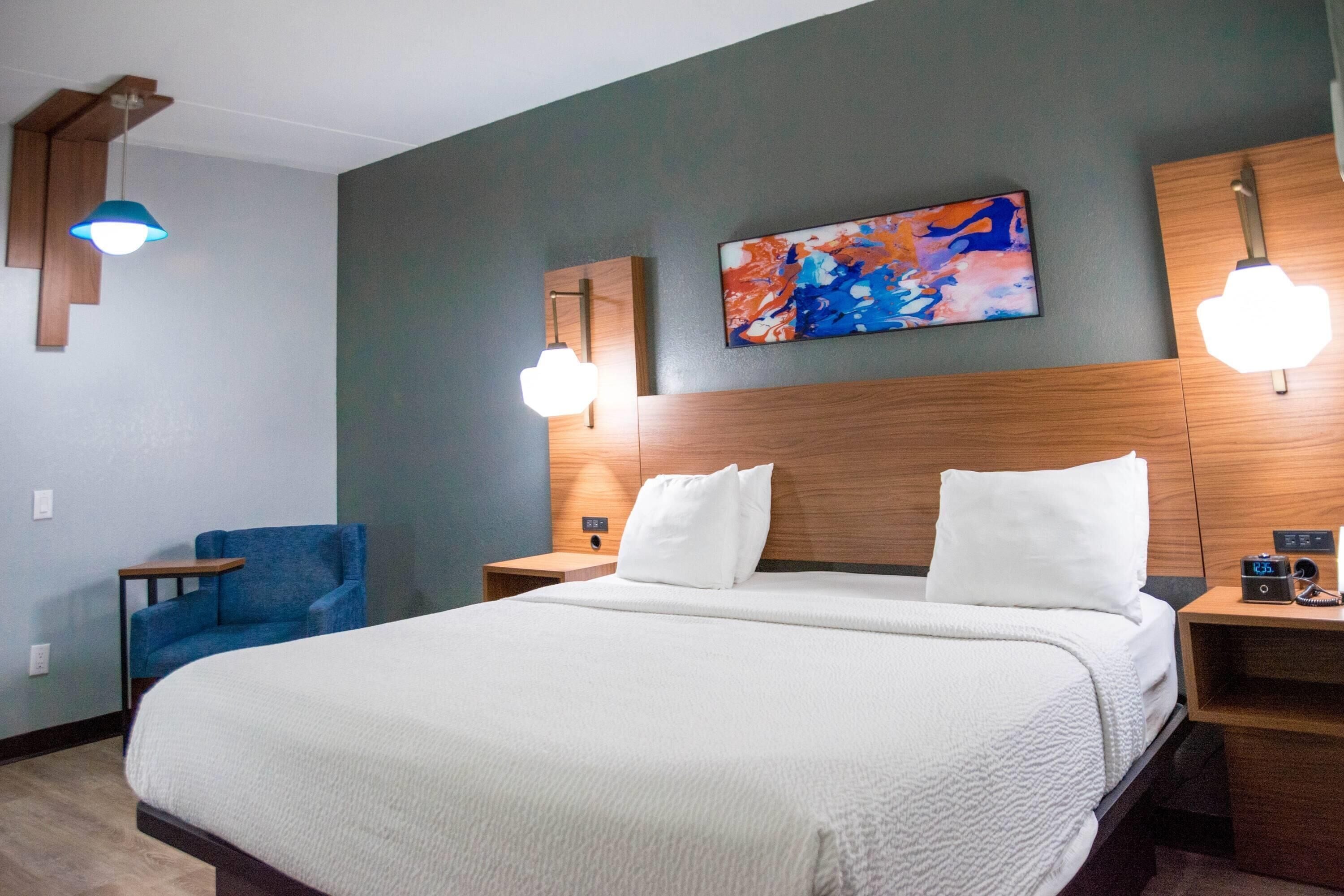 Habitación de hotel con una cama grande, un sillón azul, un cabecero de madera, un cuadro en la pared y dos lámparas de noche.