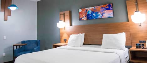 Habitación de hotel con una cama grande, un sillón azul, un cabecero de madera, un cuadro en la pared y dos lámparas de noche.