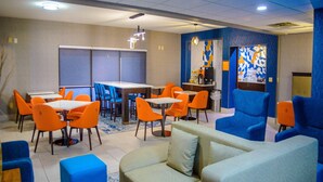 A modern dining area with orange chairs and tables, a blue sofa, and a decorative wall with abstract patterns. - La Quinta Inn & Suites by Wyndham Fayetteville I-95 (Fayetteville)