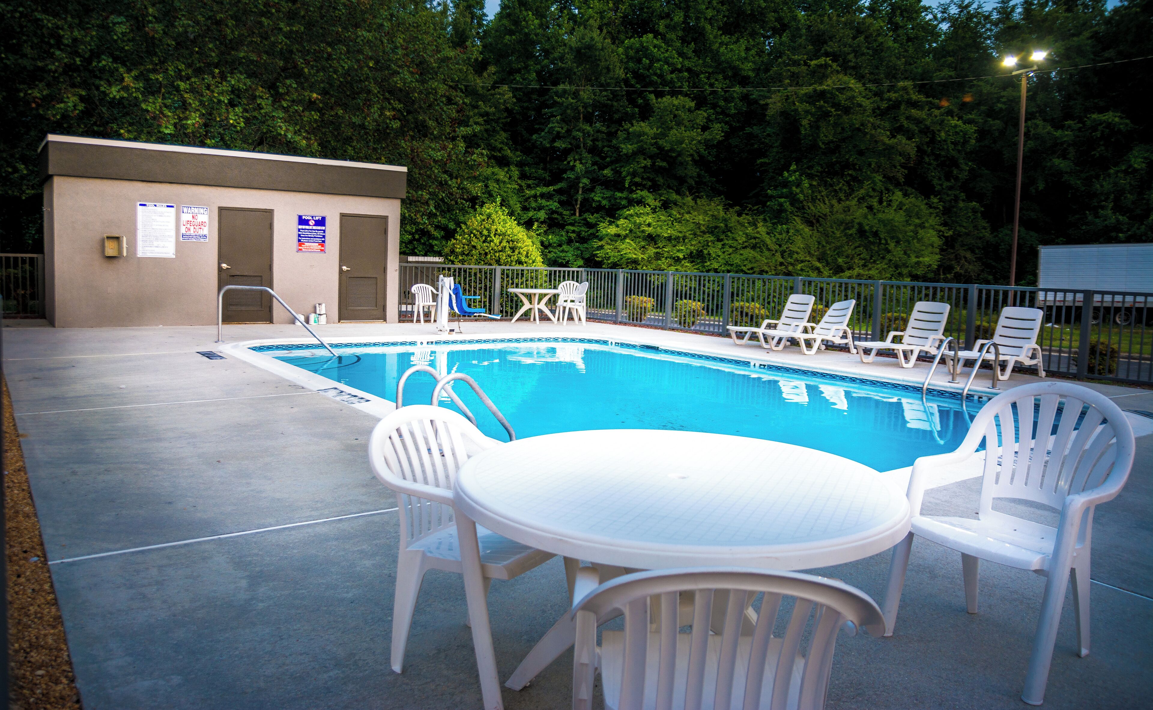 Una piscina con sillas blancas y una mesa, rodeada por una valla y árboles.