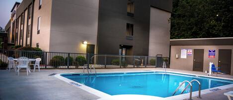 A rectangular swimming pool with a metal ladder, surrounded by a concrete deck. There are white plastic chairs and a small round table. The pool is adjacent to a building with a flat facade and multiple windows.