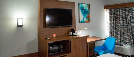 A hotel room with a bed, a desk with a flat-screen TV, a microwave, a coffee maker, a blue chair, and a window with curtains.
