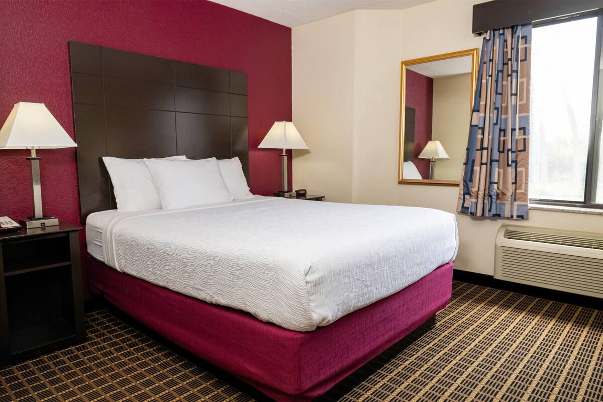 A hotel room with a large bed, two bedside tables with lamps, a large mirror, and a window with patterned curtains.