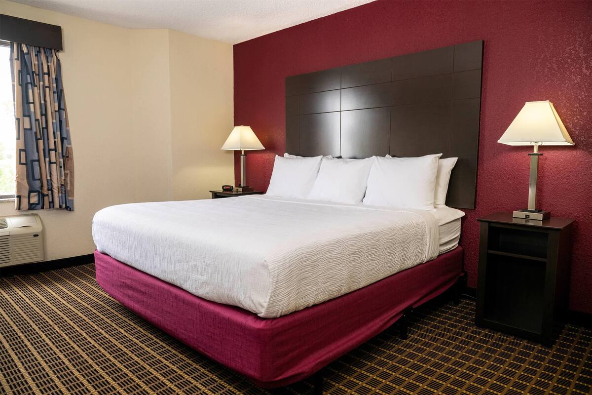 A hotel room with a large bed, two bedside tables, a window with curtains, and a red accent wall.