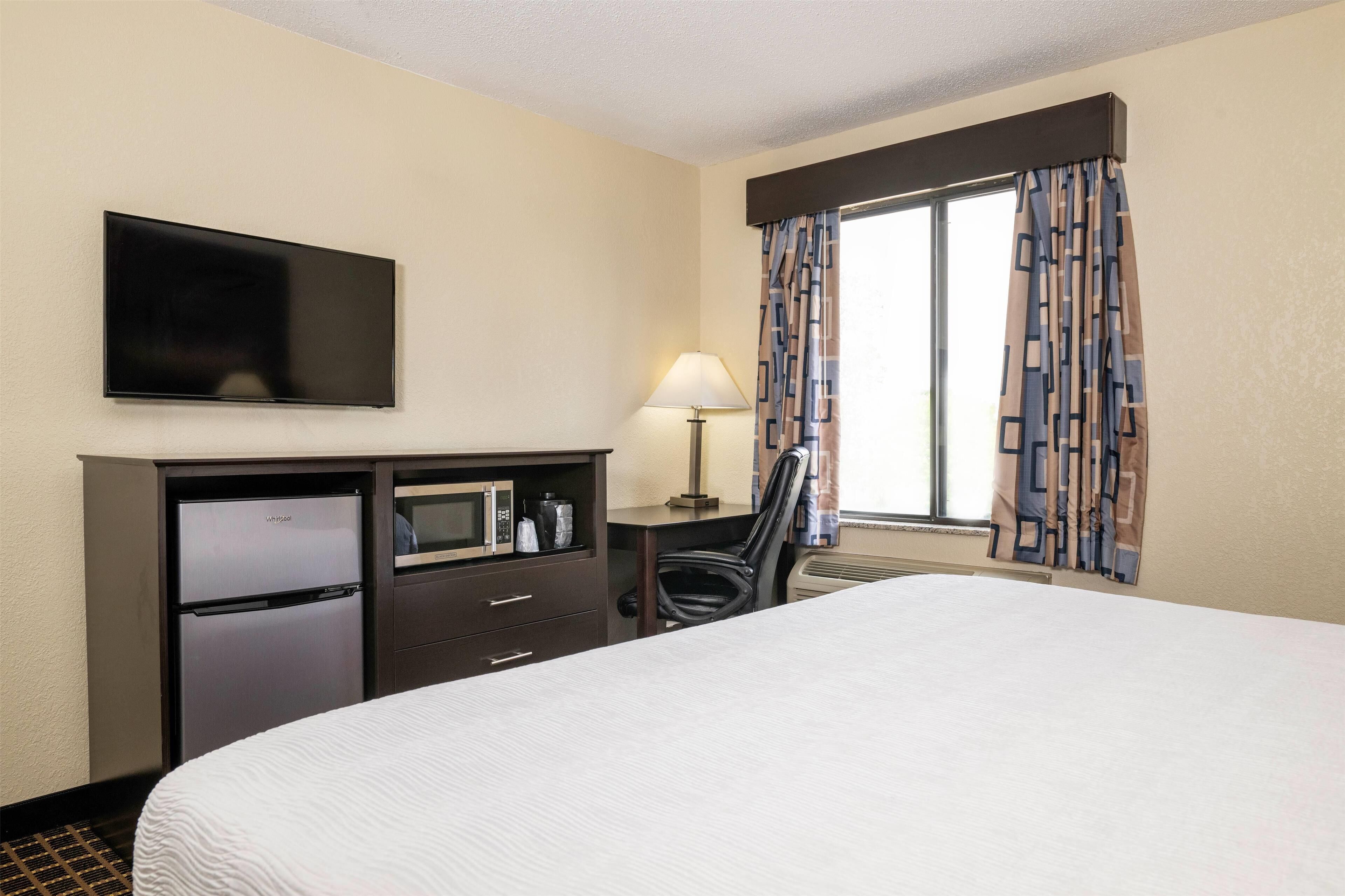 Habitación de hotel con cama, televisor de pantalla plana en la pared, minibar, escritorio con lámpara y ventana con cortinas estampadas.