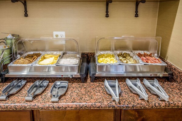 A buffet setup with two covered trays of food, including fruits, vegetables, and meats, and several serving utensils on a granite countertop.