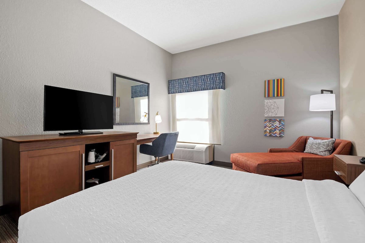 A hotel room with a large bed, a desk with a flat-screen TV, a chair, a sofa, and a nightstand with a lamp.