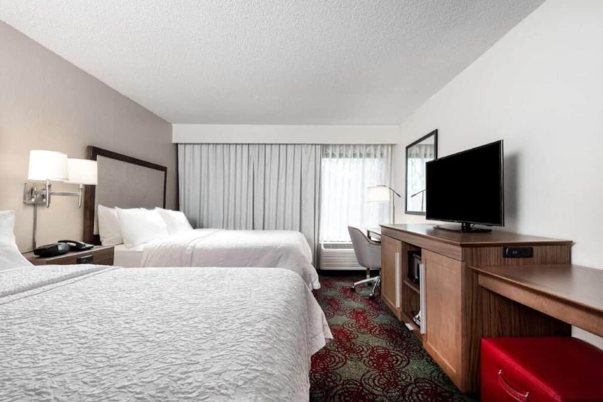 A hotel room with two beds, a desk with a television, a desk lamp, a chair, and a red bench.