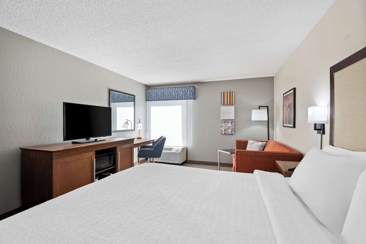 A hotel room with a large bed, a desk with a flat-screen TV, a chair, a small table, and a sofa.
