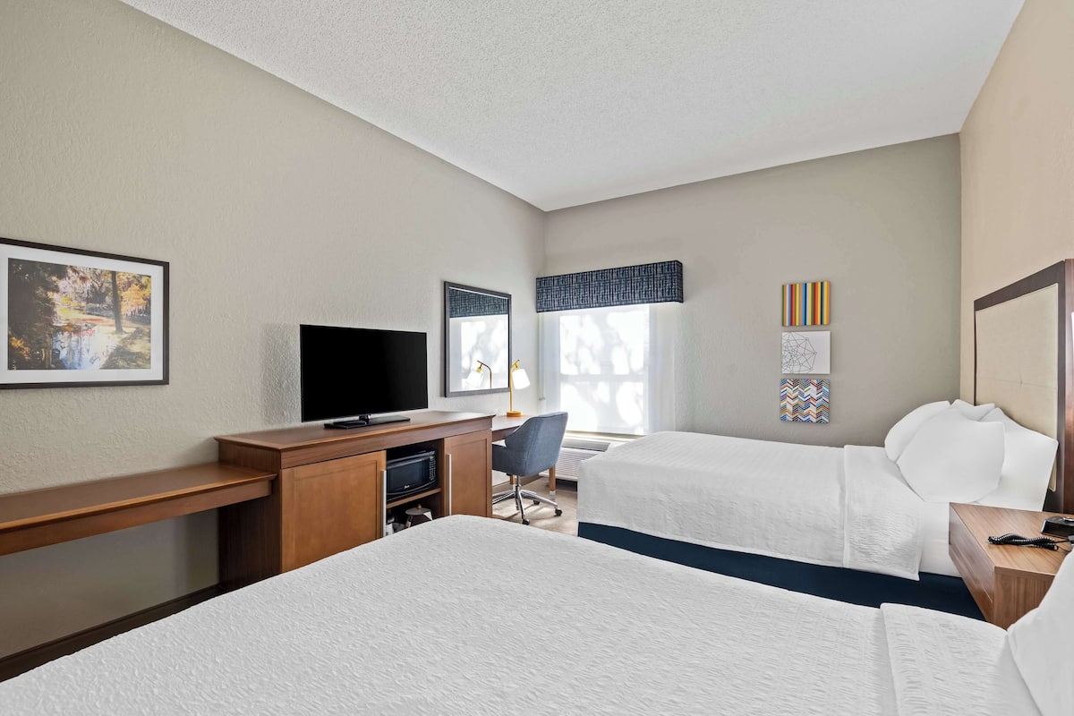 A hotel room with two beds, a desk with a flat-screen TV, a chair, and artwork on the walls.