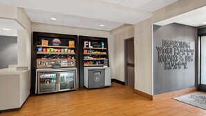 Zona d'entrada d'hotel amb sòl de fusta, dues terminals d'autoserveis, un minibar i un mur amb un missatge motivacional.