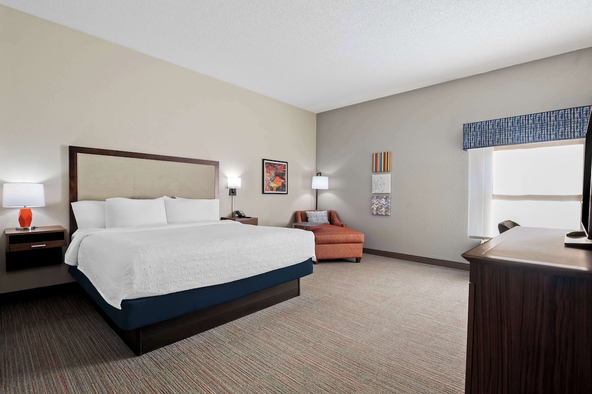 A hotel room with a large bed, a desk, a chair, a sofa, and a window with blinds.