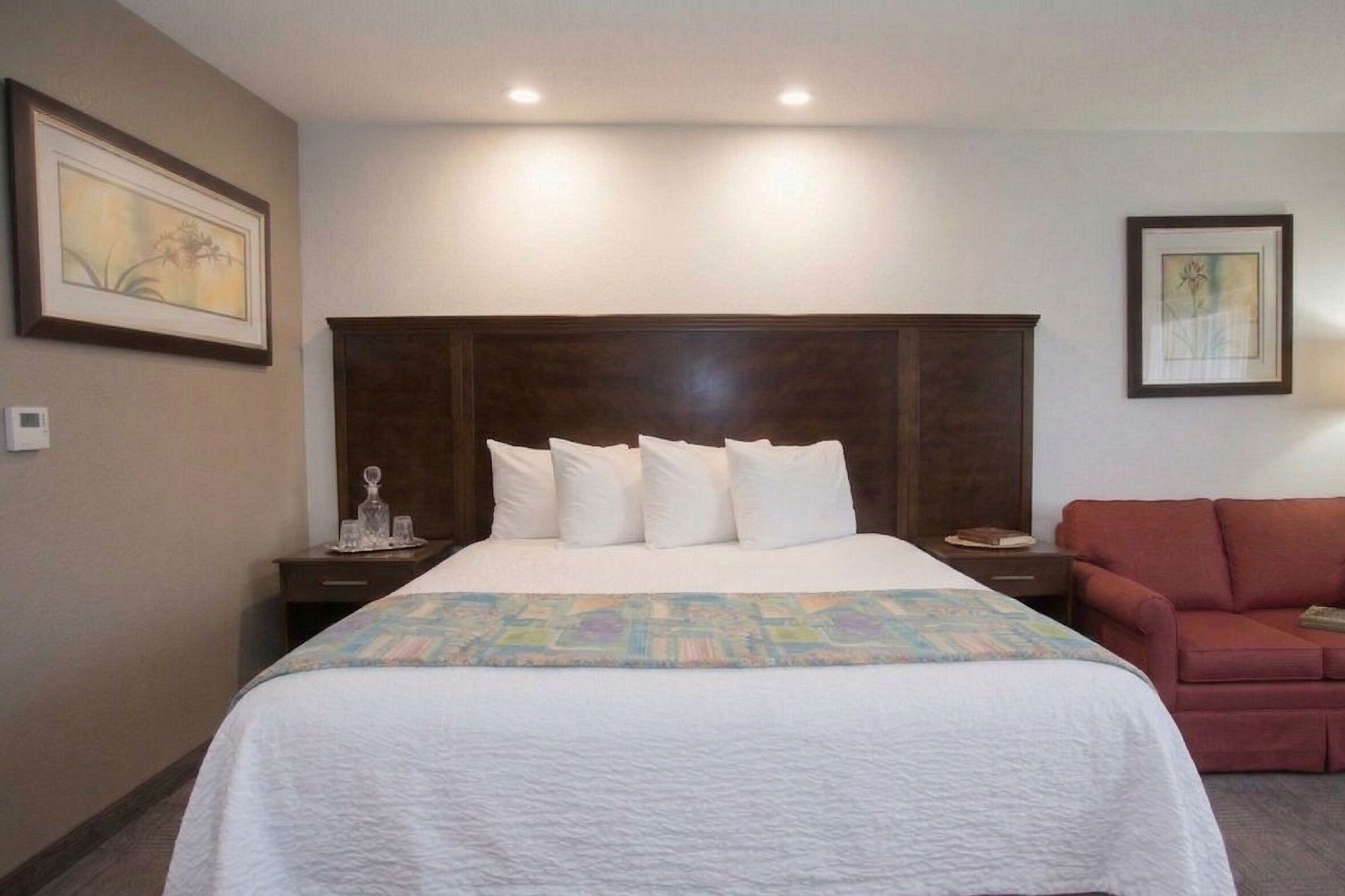 Une chambre d’hôtel avec un grand lit, une tête de lit en bois, un canapé rouge et deux tableaux encadrés accrochés aux murs.