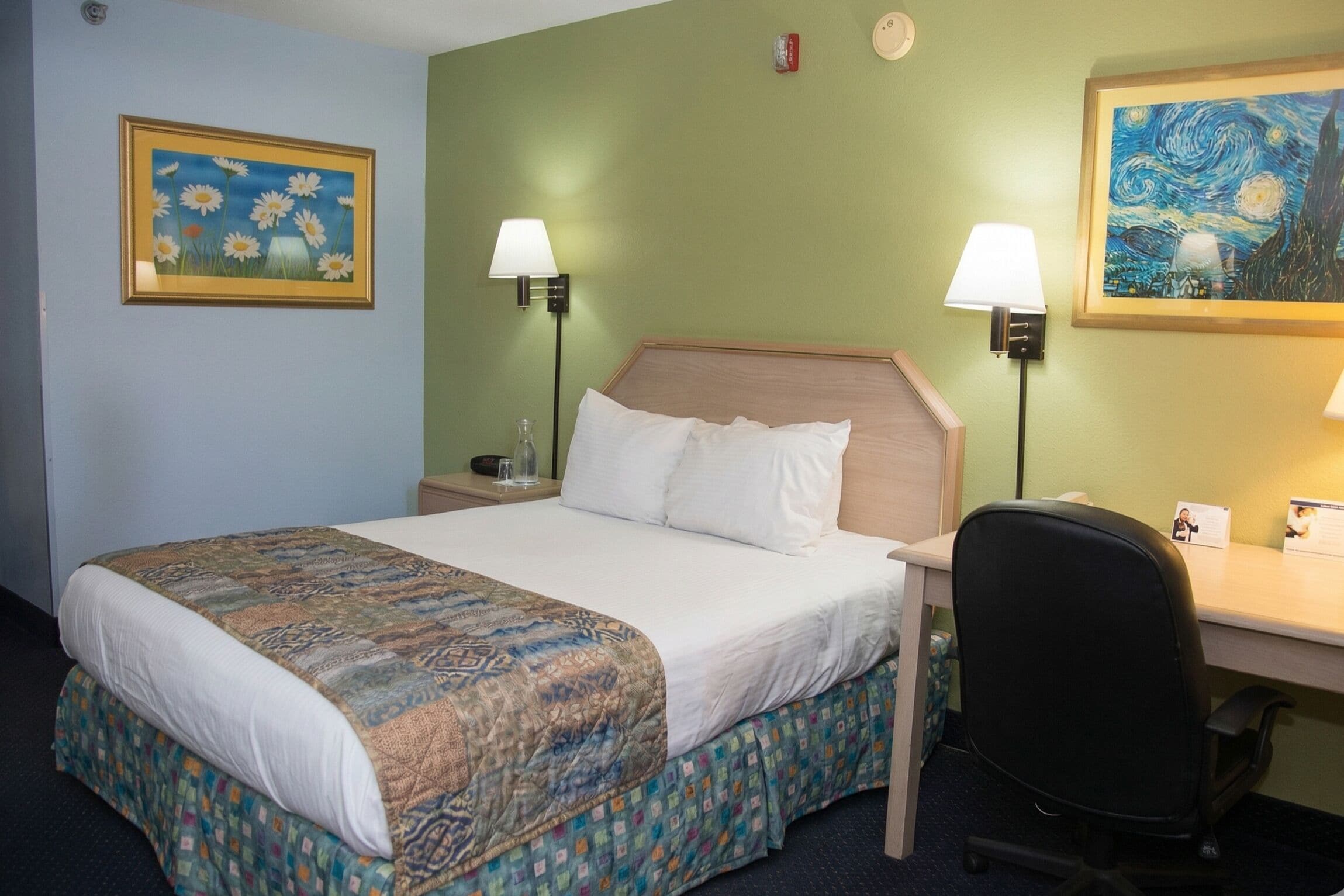 A hotel room with a bed, two bedside lamps, a desk, a chair, and two paintings on the walls.