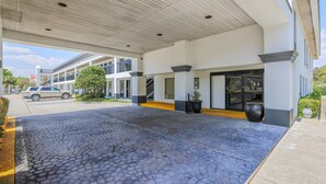 L’entrée d’un hôtel, avec une allée pavée, un espace d’attente abrité et un bâtiment doté de grandes fenêtres et d’une porte noire.
