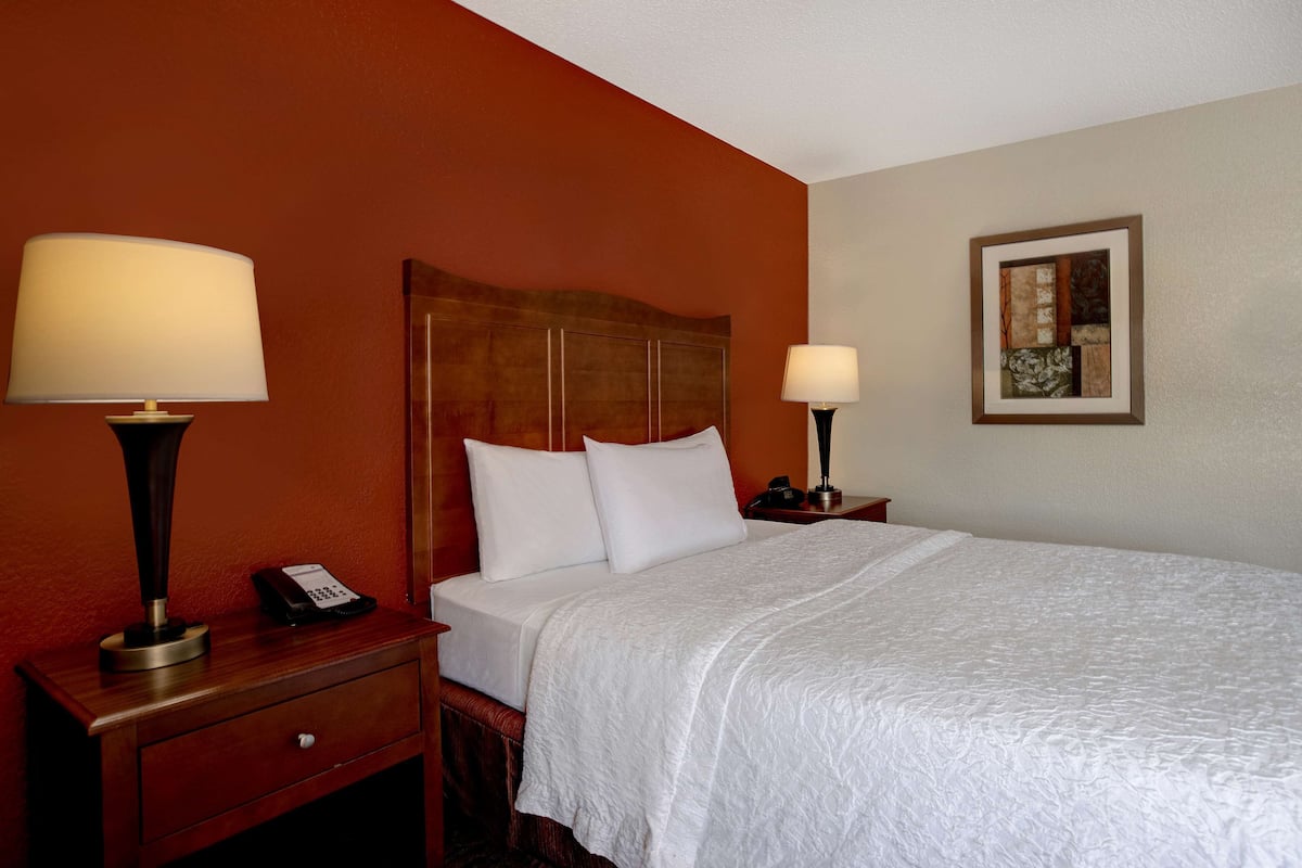A hotel room with a large bed, two bedside tables with lamps, a wooden headboard, and a framed artwork on the wall.