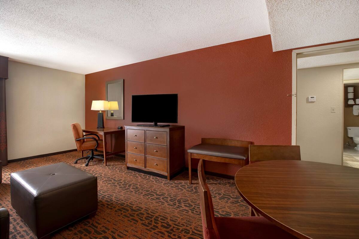 A hotel room with a large wooden dining table, a sofa, a TV on a dresser, and a patterned carpet.