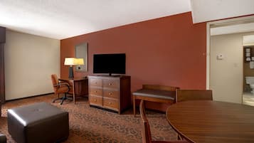 A hotel room with a large wooden dining table, a sofa, a TV on a dresser, and a patterned carpet.