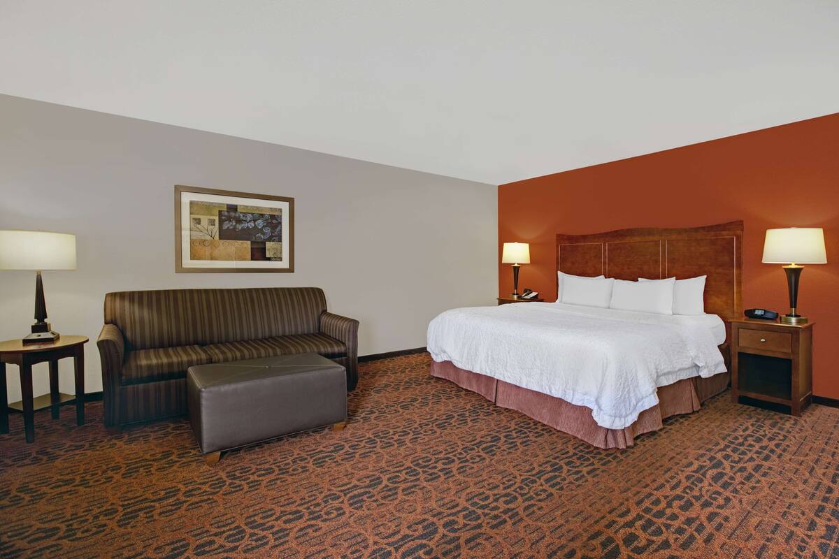 A hotel room with a bed, a sofa, an ottoman, a nightstand, a lamp, and a framed picture on the wall.