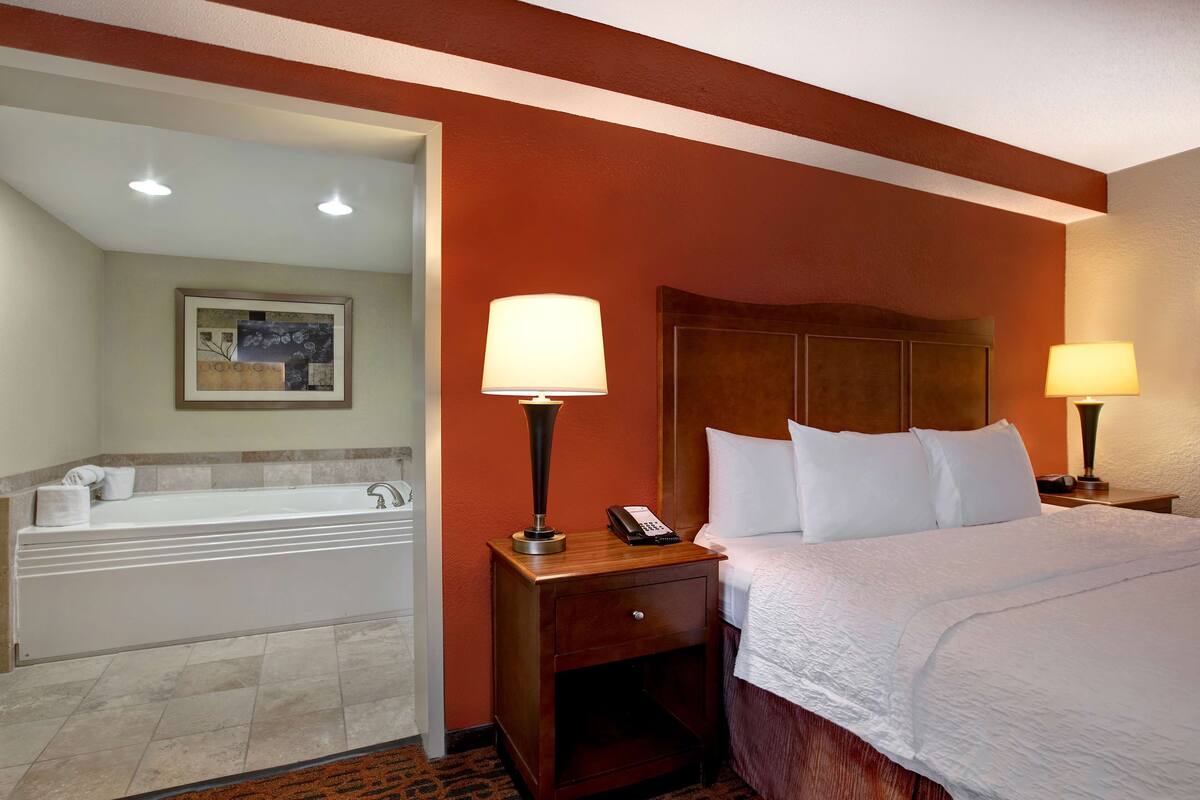 A hotel room with a large bed, two bedside lamps, a wooden nightstand, a telephone, and a bathtub.