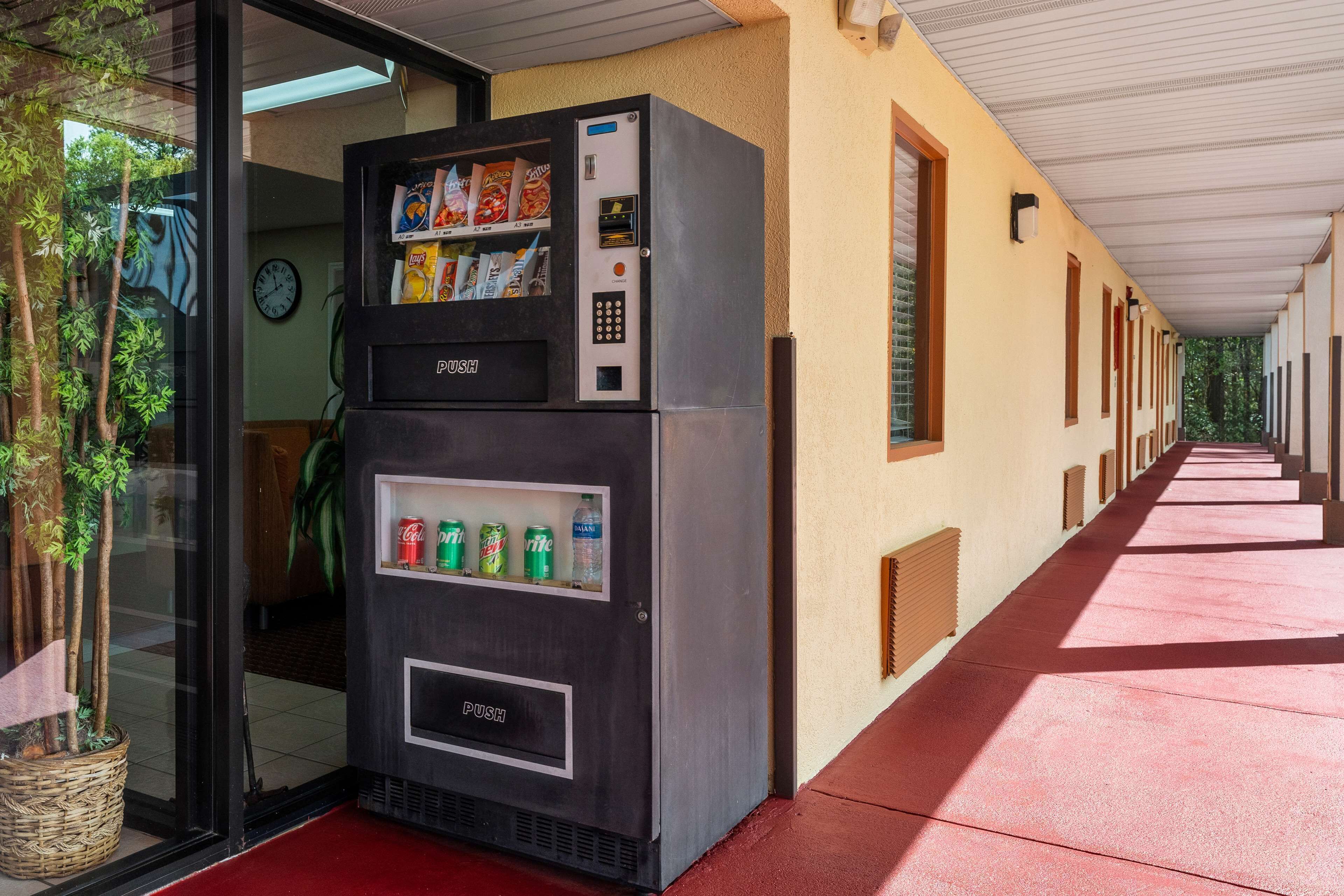 Máquina de vendas com lanches e bebidas, localizada em um corredor com carpete vermelho e um relógio na parede.