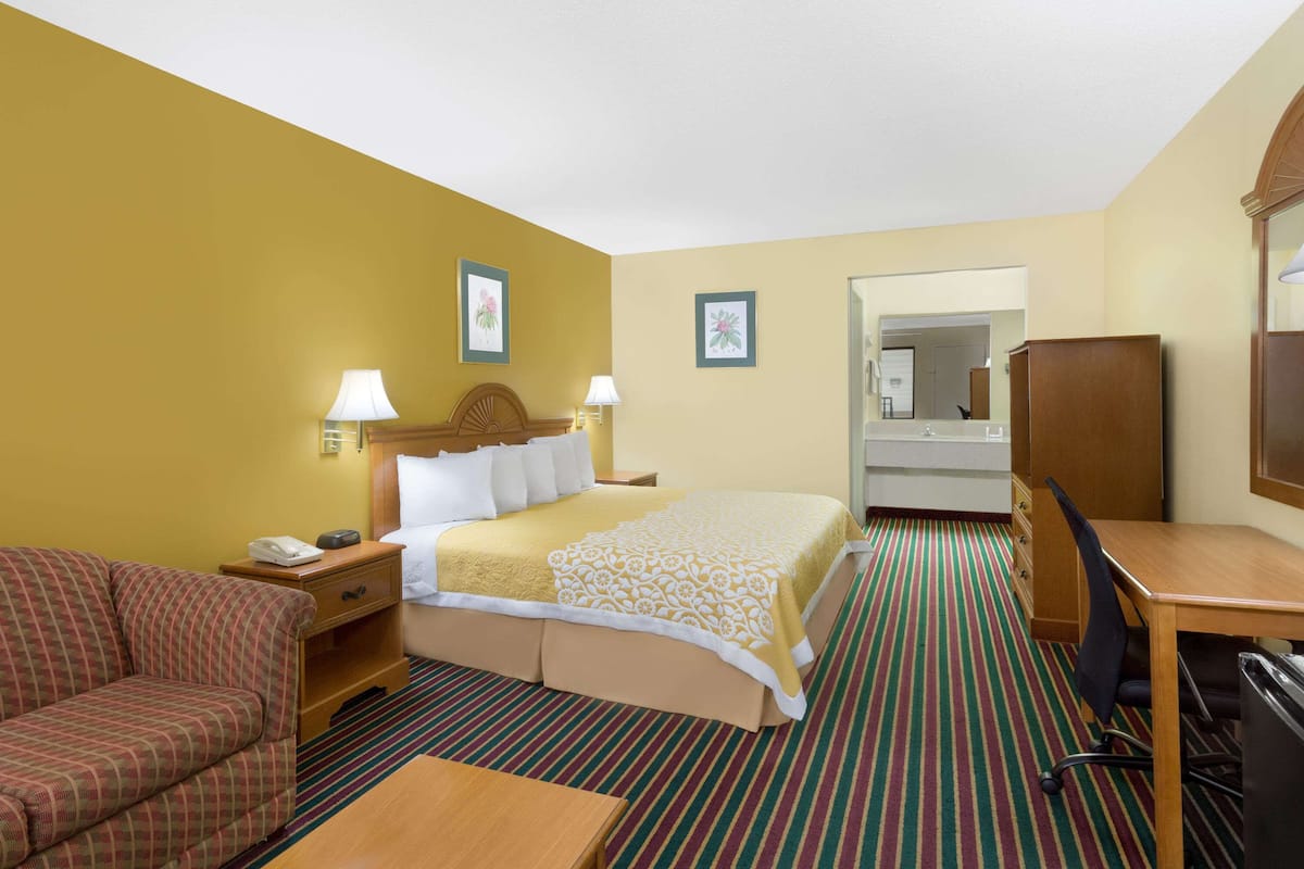 Quarto de hotel com cama, escrivaninha, poltrona, sofá e banheiro visível por uma porta aberta.